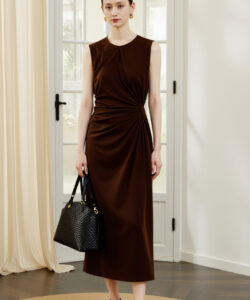 Photograph of a slender woman in a sleeveless brown dress holding a black handbag standing in a minimalist room