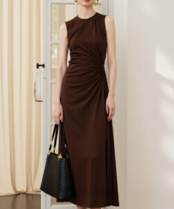 Photograph of a slender woman in a sleeveless brown dress holding a black handbag standing in a sunlit room