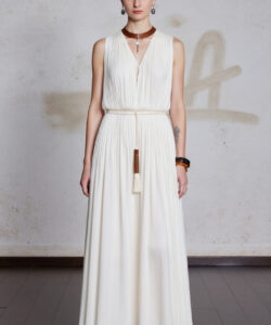 Photograph of a slender woman in a sleeveless cream pleated dress white sandals standing against a textured light gray wall