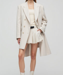 Photograph of a slender woman in oversized cream coat matching pleated skirt and white lace up boots against a plain background