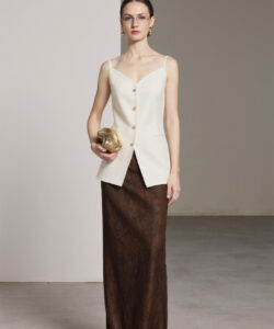 Photograph of a slender woman with glasses wearing a cream sleeveless top and brown patterned skirt holding a gold clutch