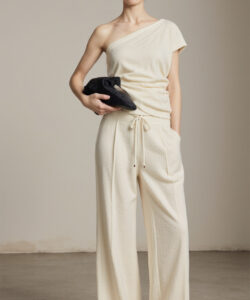 Photograph of a slender woman with light brown hair wearing a cream one shoulder top and loose pants holding a black bag