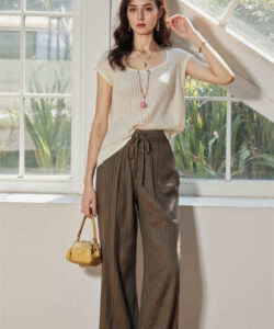 Photograph of a slender woman with long brown hair wearing a cream knit top high waisted brown pants and a yellow bag