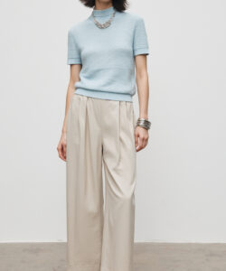 Photograph of a slender woman with short dark hair wearing a light blue turtleneck beige high waisted trousers and black sandals