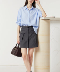 Photograph of a slim fair skinned woman in a light blue blouse gray high waisted shorts holding a brown handbag