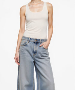Photograph of a slim fair skinned woman in a white tank top and light blue wide leg jeans against a plain background
