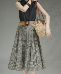 Photograph of a slim fair skinned woman in glasses wearing a sleeveless top checkered skirt and holding a woven bag