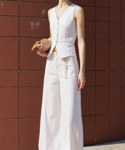 Photograph of a slim fair skinned woman in white vest and pants holding a brown clutch against a rust colored tiled wall