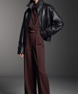 Photograph of a slim light skinned woman in a black leather jacket brown pants and brown shoes against a gray background