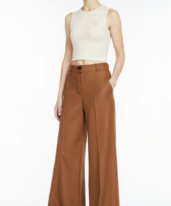 Photograph of a slim light skinned woman in a sleeveless ribbed cream top and high waisted wide legged brown trousers against a white background