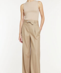 Photograph of a slim light skinned woman in beige sleeveless top and high waisted wide leg beige trousers standing against a white background