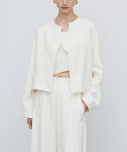 Photograph of a slim light skinned woman with dark hair in a bun wearing a cream colored oversized jacket and matching pants