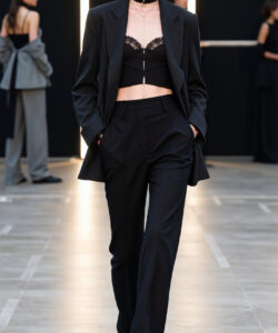 Photograph of a slim white woman in a black suit lace bralette and choker strutting confidently on a runway