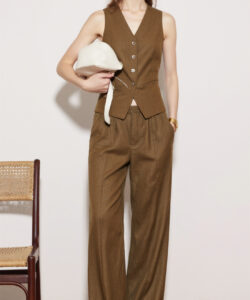 Photograph of a slim woman in a sleeveless brown vest and loose trousers holding a white hat standing against beige