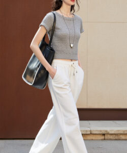 Photograph of a slim woman with dark hair wearing sunglasses a grey crop top white pants and a black bag