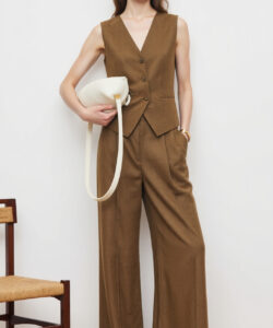 Photograph of a slim woman with long brown hair in brown vest and wide legged pants holding a white bag