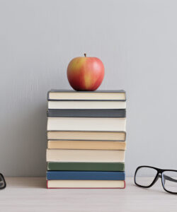Photograph-of-a-stack-of-7-books-apple-on-top-glasses-beside-against-a-plain-light-grey-background
