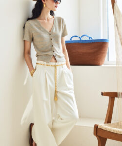 Photograph of a stylish woman in a beige hat sunglasses white pants and gold accessories leaning against a white wall