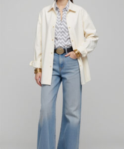 Photograph of a stylish woman in a white hat beige shirt blue jeans gold belt and accessories standing against grey