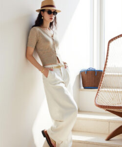 Photograph of a stylish woman in beige cardigan white pants sunglasses hat and sandals leaning on a sunlit wall