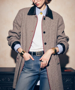 Photograph of a stylish woman in glasses black and white houndstooth coat blue jeans standing against a textured beige wall