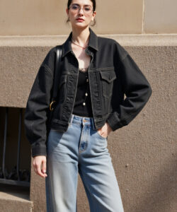 Photograph of a stylish woman in glasses black jacket blue jeans standing confidently against a beige stone wall