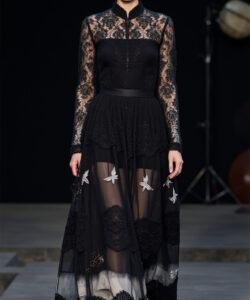 Photograph of a tall fair skinned woman in black lace dress with butterfly embroidery matching hat and gold boots runway