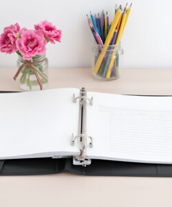 Photograph-of-a-tidy-workspace-with-a-black-binder-open-notebook-tablet-glasses-and-flower-vase-on-a-light-wood-surface