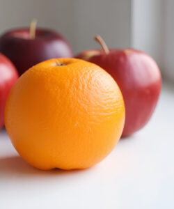 Photograph-of-a-vibrant-orange-on-a-white-surface-flanked-by-three-red-apples-near-a-window