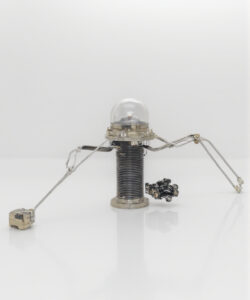 Photograph-of-a-whimsical-DIY-robot-with-a-transparent-dome-metal-body-and-small-wheels-on-a-reflective-white-surface