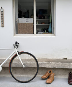 Photograph-of-a-white-bicycle-with-a-gray-basket-brown-shoes-and-a-brown-leather-handbag-in-front-of-a-white-wall