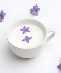 Photograph-of-a-white-ceramic-cup-filled-with-milk-adorned-with-scattered-purple-flowers-on-a-clean-white-background