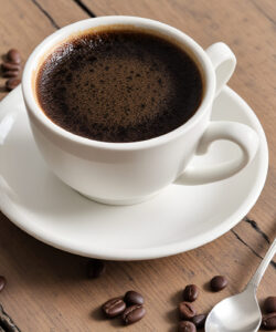 Photograph-of-a-white-ceramic-cup-of-dark-coffee-on-a-saucer-surrounded-by-scattered-coffee-beans-on-a-wooden-table