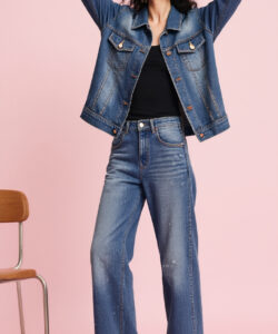 Photograph of a woman in a blue denim jacket and high waisted jeans standing against a pink background arms raised