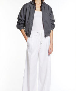 Photograph of a woman in a gray zip up jacket white tank top white pants and black sandals standing against white