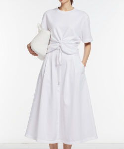 Photograph of a woman in a white short sleeved dress with a knot holding a white bag standing against a white background