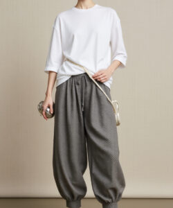Photograph of a woman in a white t shirt grey loose pants white flats and glasses against a beige background