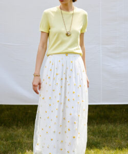 Photograph of a woman in a yellow top white floral skirt lace headband gold jewelry standing on grass