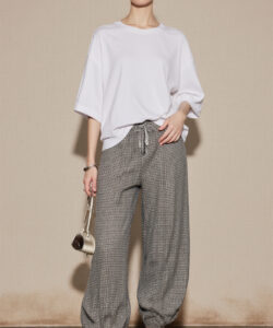 Photograph of a woman in white top patterned pants white shoes glasses and clutch standing against a beige textured wall