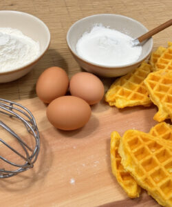 Photograph-of-a-wooden-table-with-a-bowl-of-flour-eggs-a-waffle-iron-and-waffle-batter-on-a-plate