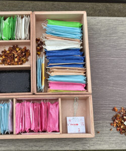 Photograph-of-a-wooden-tea-box-with-colorful-tea-packets-tea-leaves-and-a-metal-tea-infuser-on-a-table