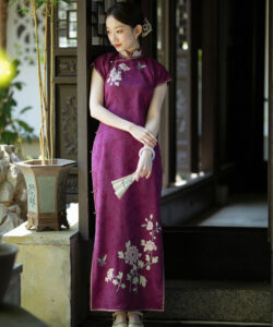 Photograph of a young asian woman in a maroon cheongsam with floral embroidery holding a fan standing in a traditional setting