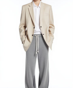 Photograph of a young curly haired man in glasses beige blazer white shirt gray pants and slippers against a plain white background