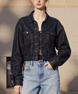 Photograph of a young slim woman with light skin brown hair and glasses wearing a black denim jacket and jeans