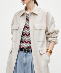 Photograph of a young woman in a beige trench coat striped shirt high waisted jeans wide brim hat gold jewelry neutral background