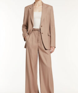 Photograph of a young woman in a light brown suit white tank top and beige high heeled sandals standing against white