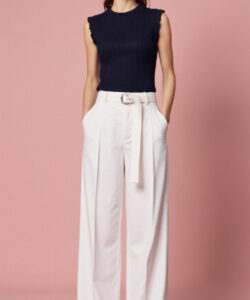 Photograph of a young woman in a sleeveless navy top and high waisted white pants against a solid pink background