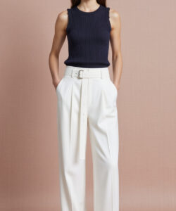 Photograph of a young woman in a sleeveless navy top white high waisted pants and black sandals against a beige backdrop