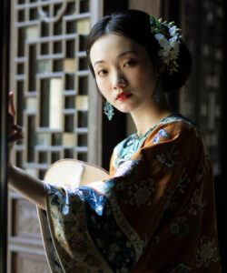 Photograph of an asian woman in traditional chinese attire adorned with flowers standing by a wooden lattice window
