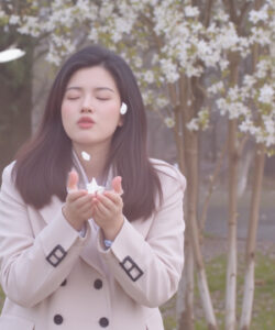 Photograph-of-an-Asian-woman-with-long-dark-hair-wearing-a-beige-coat-blowing-white-petals-in-a-spring-park